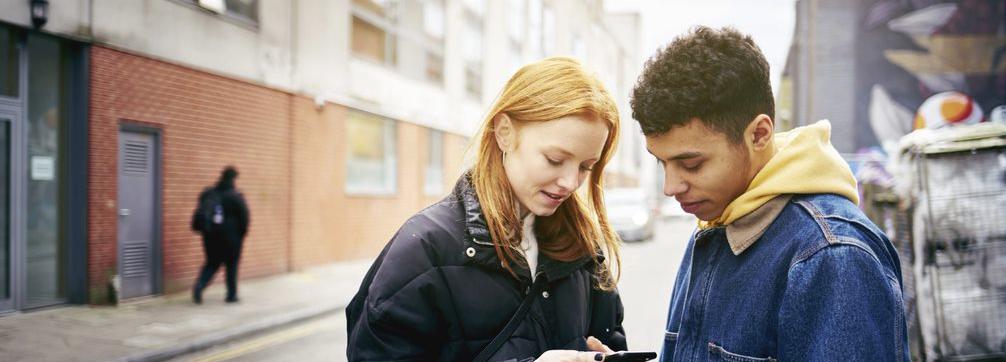 Woman and man standing in a street both looking at her phone.