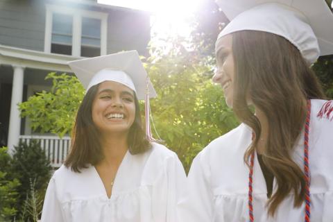 Two women wearing graduation gowns and mortar boards smiling at each other.